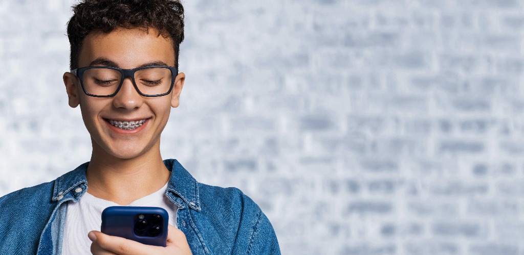 Lächelnd schwarzes lockiges Haar lustiger junger Mann, Kerl, Junge tragen Zahnspange halten Hand tippen Text, Blick lesen Blick auf Handy mobile Smartphone Handy, stehen gegen weiße Ziegel Loft Wand Hintergrund